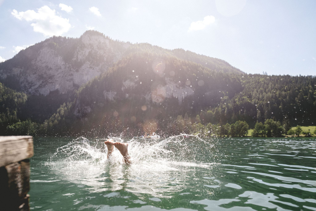 Person springt kopfüber in klaren Bergsee vor bewaldeter Alpenkulisse